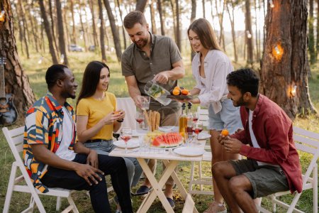 Téléchargez les photos : Réunion d'amis multiraciaux dînant et buvant du vin pendant la fête dans la forêt - en image libre de droit
