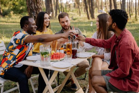 Photo for Meeting of multiracial group of friends eating dinner and drinking wine during party in the forest - Royalty Free Image