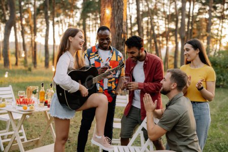 Téléchargez les photos : Attrayant hipster femme jouer de la guitare gagner sur les hommes au dîner avec des amis multiraciaux - en image libre de droit