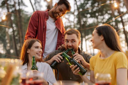 Foto de Amigos multirraciales divirtiéndose en una fiesta, chicos y chicas bromeando mientras abren cerveza con sus dientes - Imagen libre de derechos