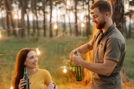 Foto de Barbudo hombre hipster abre una cerveza en una fiesta con amigos en un bosque decorado con lámparas colgantes - Imagen libre de derechos