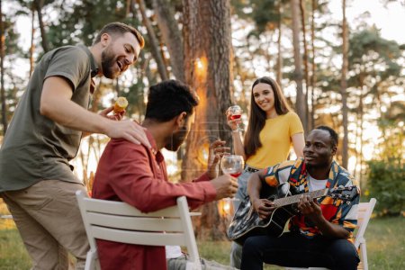 Foto de Grupo multirracial de amigos, hombre africano tocando la guitarra alrededor de amigos felices bebiendo vino - Imagen libre de derechos