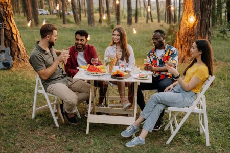 Photo for Multiracial group of happy friends sitting around table drinking wine talking at dinner party in evening summer forest - Royalty Free Image