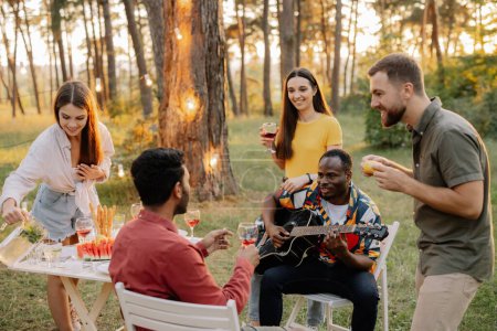 Foto de Grupo multirracial de amigos, hombre africano tocando la guitarra alrededor de amigos felices bebiendo vino - Imagen libre de derechos