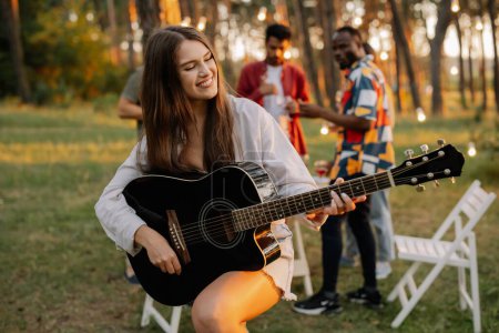 Foto de Hermosa chica hipster tocando la guitarra en el fondo de una cena al aire libre de amigos multirraciales - Imagen libre de derechos