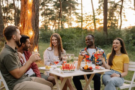 Foto de Grupo multirracial de amigos felices sentados alrededor de la mesa bebiendo vino hablando en la cena en el bosque de verano por la noche - Imagen libre de derechos