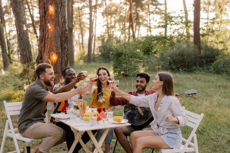 Téléchargez les photos : Rencontre d'amis multiraciaux jouant de la guitare, chantant, dînant et buvant du vin pendant la fête dans la forêt - en image libre de droit
