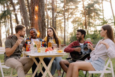 Photo for Meeting of multiracial group of friends playing guitar, singing, eating dinner and drinking wine during party in the forest - Royalty Free Image