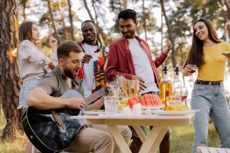 Foto de Grupo multicultural de personas, hombre hipster barbudo tocando la guitarra y amigos bailando y divirtiéndose - Imagen libre de derechos