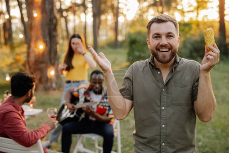 Photo for Bearded hipster man eating corn on the background of dinner party of multiracial friends - Royalty Free Image