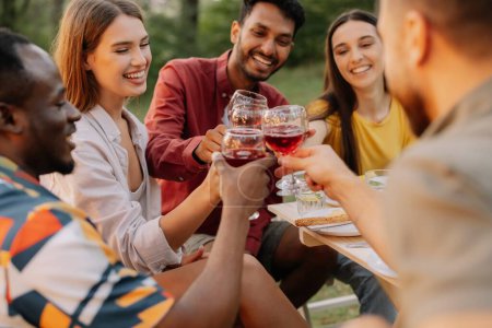 Téléchargez les photos : Groupe multiracial d'amis heureux assis autour de la table buvant du vin lors d'un dîner dans la forêt d'été du soir - en image libre de droit