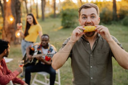 Photo for Bearded hipster man eating corn on the background of dinner party of multiracial friends - Royalty Free Image