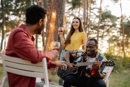 Téléchargez les photos : Groupe multiracial d'amis, homme africain jouant de la guitare autour d'amis heureux buvant du vin - en image libre de droit