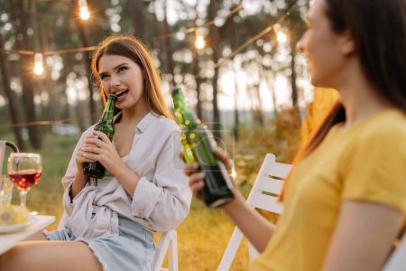 Téléchargez les photos : Funny hipster femme ouvre la bière avec des dents à la fête avec des amis dans la forêt décorée avec des lampes suspendues - en image libre de droit