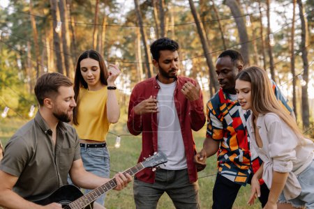 Foto de Grupo multirracial de personas, hipster barbudo tocando la guitarra y amigos bailando, cantando y divirtiéndose - Imagen libre de derechos