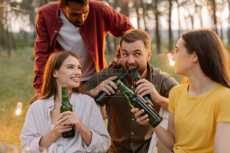 Téléchargez les photos : Amis multiraciaux s'amusant à une fête, les garçons et les filles plaisantent tout en ouvrant la bière avec leurs dents - en image libre de droit