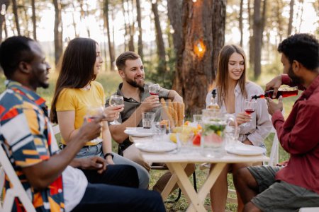 Photo for Meeting of multiracial group of friends eating dinner and drinking wine during party in the forest - Royalty Free Image