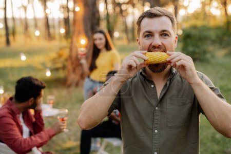 Téléchargez les photos : Homme hipster barbu mangeant du maïs sur le fond du dîner d'amis multiraciaux - en image libre de droit