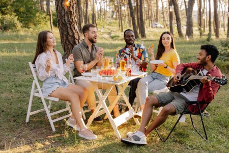 Foto de Encuentro de grupo multirracial de amigos tocando la guitarra, cantando, cenando y bebiendo vino durante la fiesta en el bosque - Imagen libre de derechos