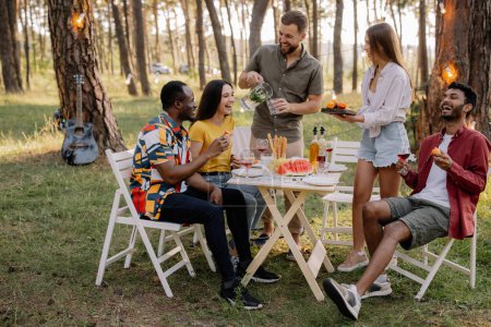 Foto de Reunión del grupo multirracial de amigos cenando y bebiendo vino durante la fiesta en el bosque - Imagen libre de derechos