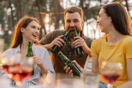 Téléchargez les photos : Amis multiraciaux s'amusant à une fête, les garçons et les filles plaisantent tout en ouvrant la bière avec leurs dents - en image libre de droit