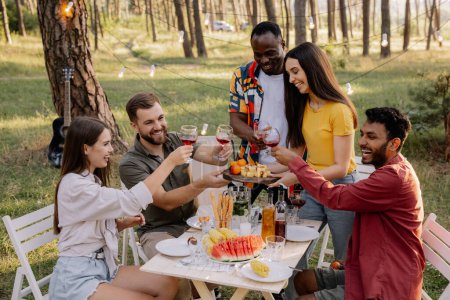 Foto de Reunión del grupo multirracial de amigos cenando y bebiendo vino durante la fiesta en el bosque - Imagen libre de derechos