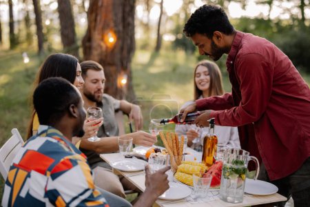 Téléchargez les photos : Réunion d'amis multiraciaux dînant et buvant du vin pendant la fête dans la forêt - en image libre de droit