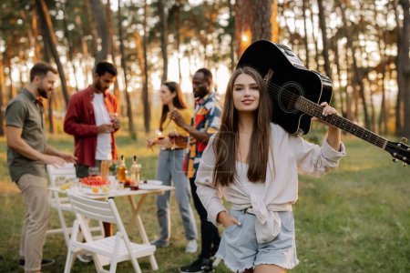 Photo for Beautiful hipster girl playing the guitar on the background of a dinner party outdoors of multiracial friends - Royalty Free Image