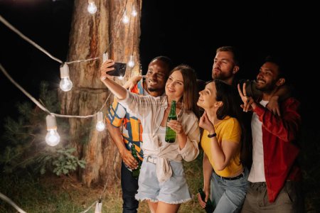 Foto de Un grupo de amigos multirraciales divirtiéndose y bebiendo cerveza se toman selfies cerca de lámparas colgantes por la noche - Imagen libre de derechos