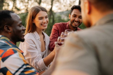 Photo for Multiracial group of happy friends sitting around table drinking wine talking at dinner party in evening summer forest - Royalty Free Image