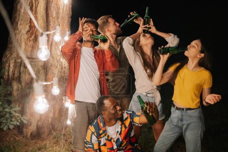 Photo for A company of multiracial friends drinking beer at party, making faces near hanging lamps - Royalty Free Image