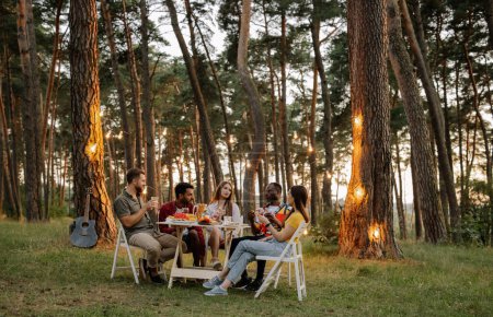 Foto de Grupo multirracial de amigos felices sentados alrededor de la mesa bebiendo vino hablando en la cena en el bosque de verano por la noche - Imagen libre de derechos