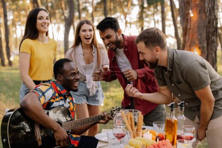 Foto de Grupo multicultural de personas, hipster africano tocando la guitarra y amigos bailando, cantando y divirtiéndose - Imagen libre de derechos