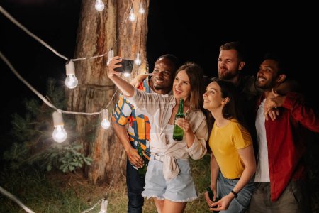 Téléchargez les photos : Un groupe d'amis multiraciaux s'amuser et boire de la bière prennent selfies près des lampes suspendues la nuit - en image libre de droit