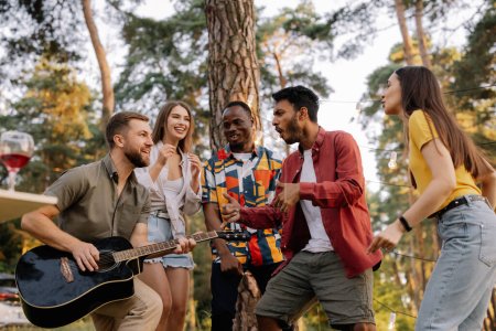 Photo for Multiracial group of people, bearded hipster man playing guitar and friends dancing, singing and having fun - Royalty Free Image
