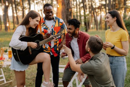 Téléchargez les photos : Attrayant hipster femme jouer de la guitare gagner sur les hommes au dîner avec des amis multiraciaux - en image libre de droit