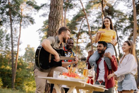 Foto de Grupo multicultural de personas, hipster barbudo tocando la guitarra y amigos bailando, cantando y divirtiéndose - Imagen libre de derechos