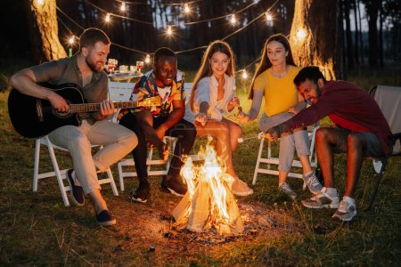 Photo for Group of multiracial restful friends roasting marshmallows while sitting by bonfire - Royalty Free Image