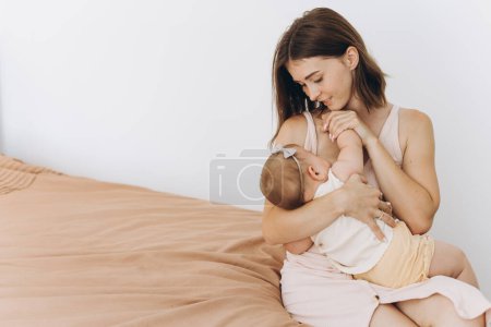 Photo for Happy mother breastfeeding a baby girl on a bed in a bright room - Royalty Free Image