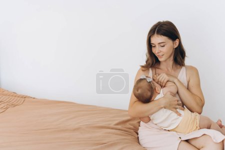 Foto de Madre feliz amamantando a una niña en una cama en una habitación luminosa - Imagen libre de derechos