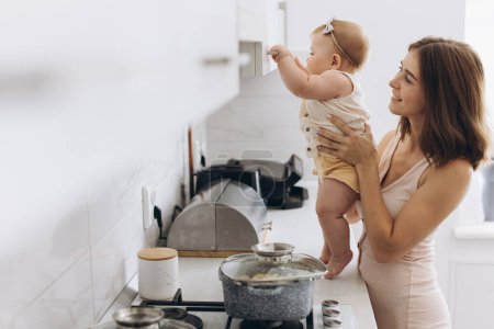 A mother with her little baby daughter prepares food in the kitchen