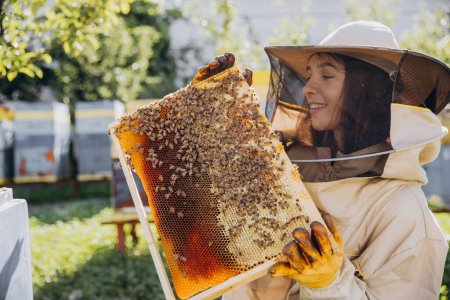 Feliz hembra apicultora sonriente en traje protector sosteniendo marco de abeja con abejas en el colmenar