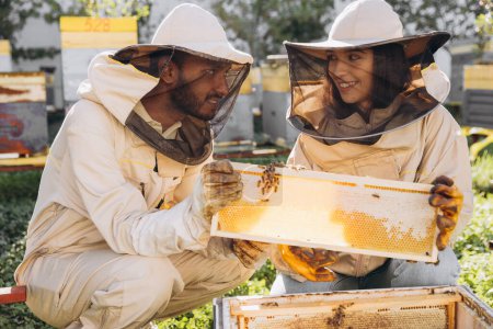 Téléchargez les photos : Couple d'apiculteurs souriants heureux travaillant avec des outils apicoles près de ruche à la ferme apicole - en image libre de droit