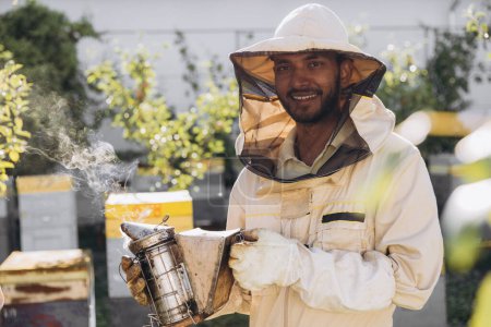 Téléchargez les photos : Apiculteur indien souriant heureux fumant des abeilles mellifères avec fumeur d'abeilles sur le rucher - en image libre de droit