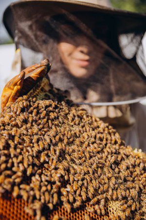Primer plano hembra apicultora en traje protector que sostiene el marco de abejas con abejas en el colmenar