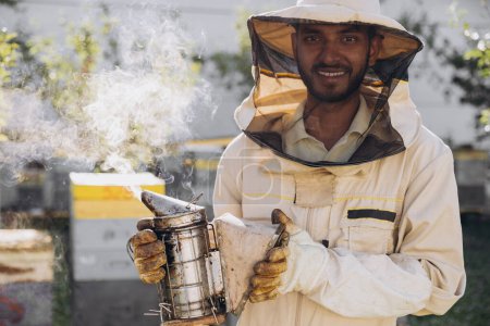 Foto de Feliz sonriente macho indio apicultor fumando abejas con abeja fumador en el colmenar - Imagen libre de derechos
