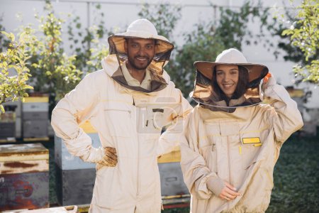 Téléchargez les photos : Deux apiculteurs en uniforme de protection debout ensemble près des ruches en bois sur un petit rucher traditionnel - en image libre de droit