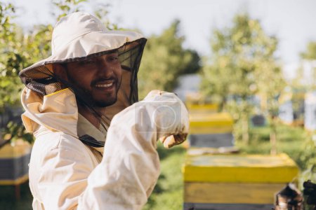 Téléchargez les photos : Joyeux apiculteur indien souriant en uniforme debout dans un rucher et tenant une abeille dans une ferme apicole - en image libre de droit
