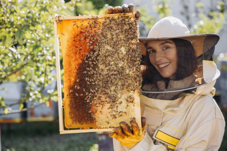Foto de Feliz hembra apicultora sonriente en uniforme de pie en colmenar y con marco de abeja - Imagen libre de derechos