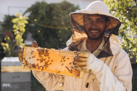 Téléchargez les photos : Heureux apiculteur indien souriant dans un uniforme debout dans le rucher et tenant un cadre d'abeille à la ferme d'abeilles - en image libre de droit